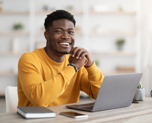 Positive black man freelancer sitting at workdesk at home, using laptop, happy african american guy enjoying remote job, sending emails to clients or surfing on Internet, smiling at camera, copy space