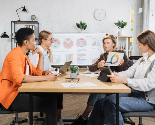 Caucasian mature woman in suit showing financial statistic of enterprise on glass board during working meeting with diverse females colleagues. Cooperation, technology and people concept.