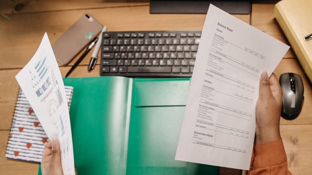 Two hands holding printed financial statements over a wooden desk with a keyboard, a mouse and a green folder