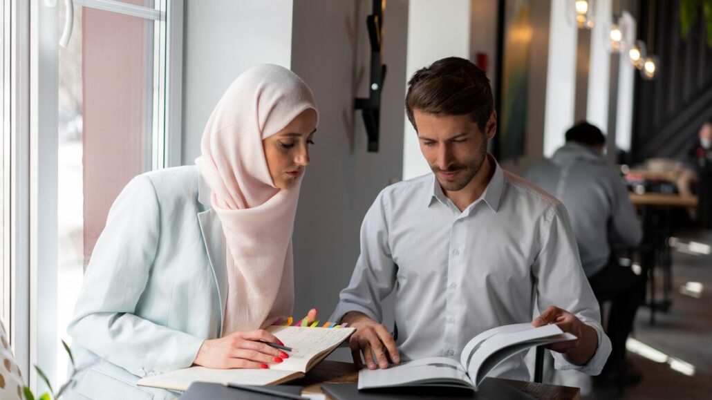 A woman in a headscarf and a man in a shirt reading through papers and a notebook together at a table by a bright window