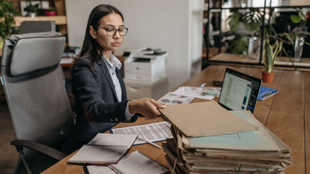 Woman in glasses at a wooden office desk sorting a stack of paper files beside a laptop showing a spreadsheet