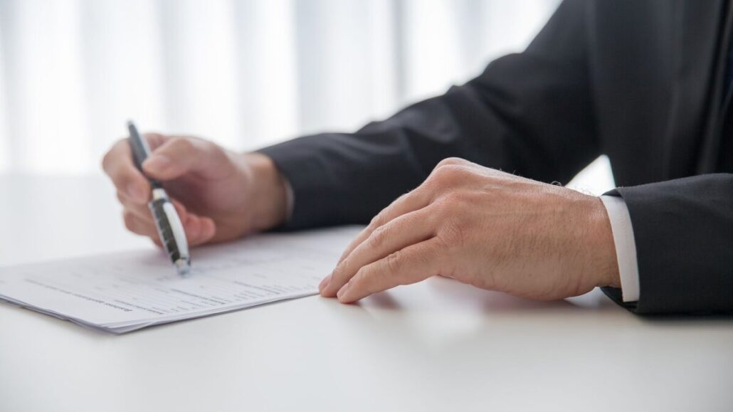 A person in a dark suit signing a printed document with a pen at a white desk