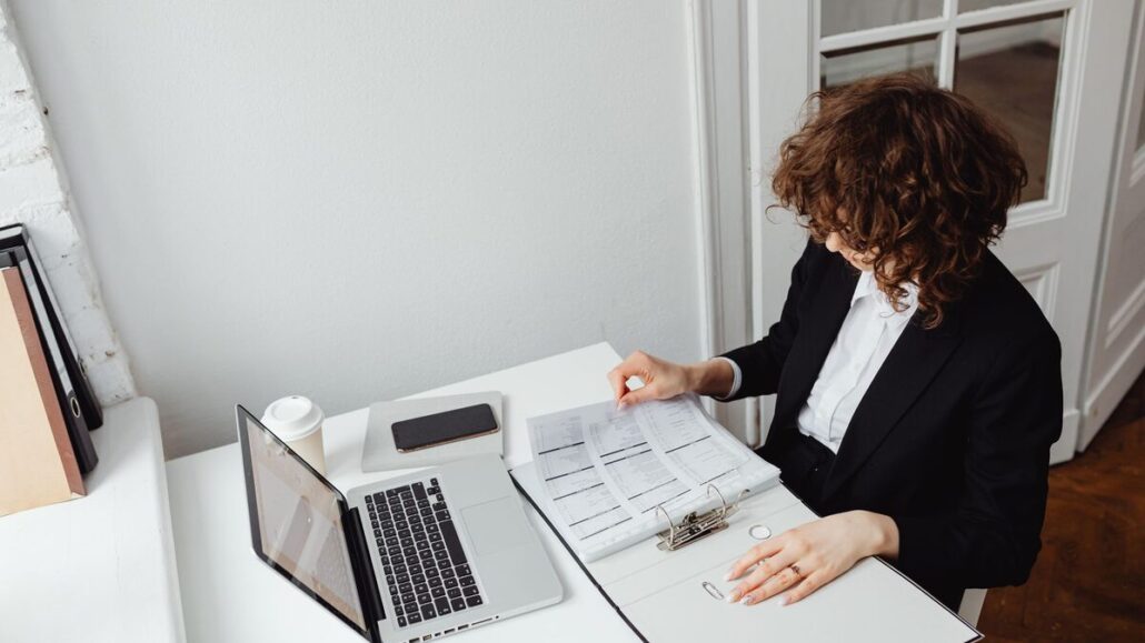 A woman in a black blazer working at a white desk by a window, checking printed documents beside an open laptop and phone