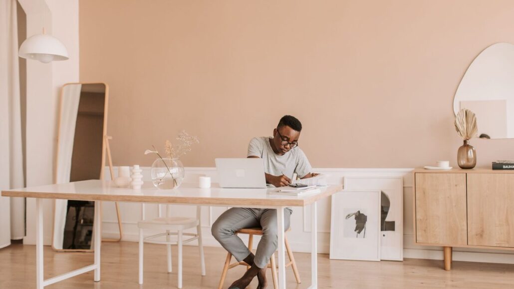 A man working at a laptop and writing in a notebook at a long white table in a bright, minimal home office