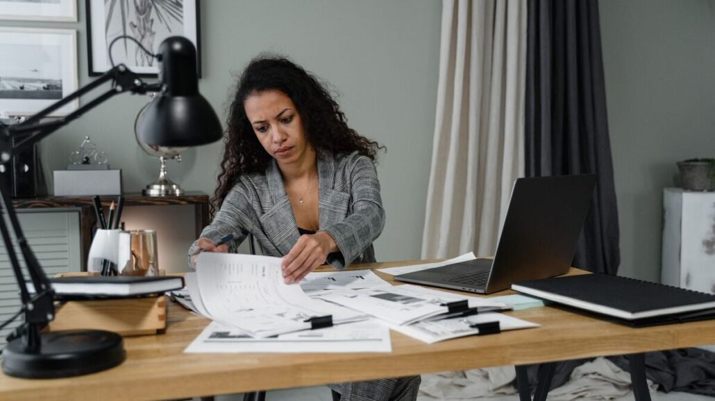 A woman with curly hair reading printed documents at a wooden desk with a desk lamp, notebooks and a laptop