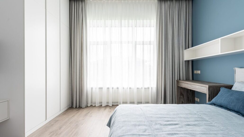A bright, tidy bedroom with a made double bed, a large window with sheer curtains and a blue feature wall