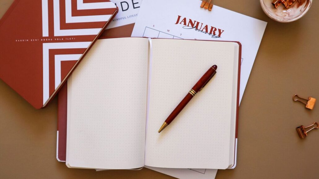A flat lay of an open notebook with a red pen on a brown desk, beside a January calendar page and paper clips