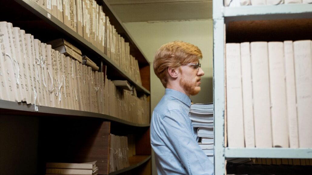 A man in a light blue shirt placing a stack of files on a shelf in a narrow archive room lined with document boxes