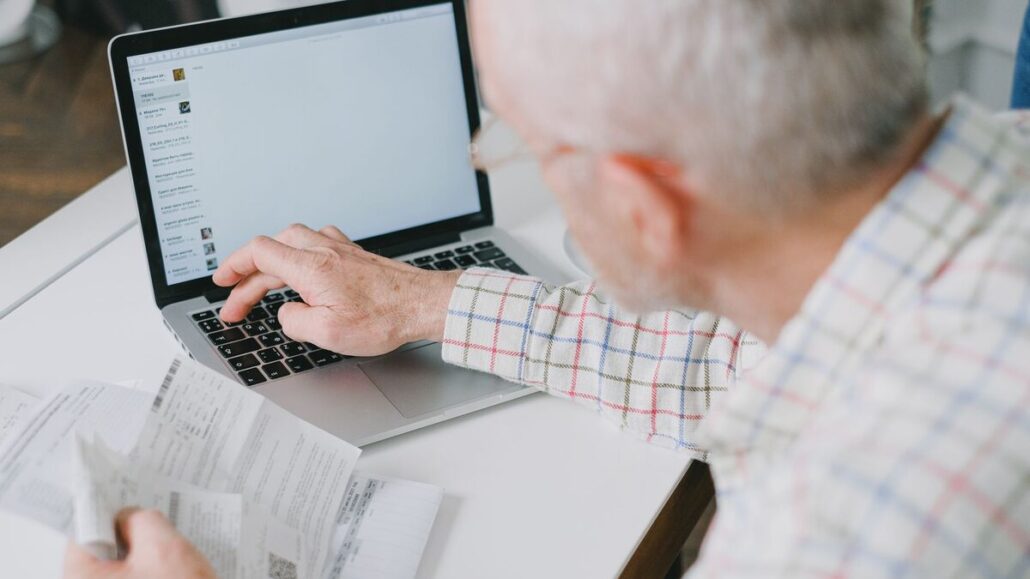 An older man in a checked shirt typing on a laptop at a white desk while holding a printed paper document