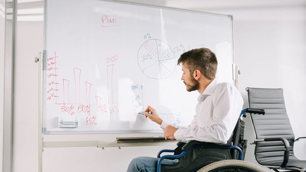 A man using a wheelchair drawing a bar chart on a whiteboard in a bright office
