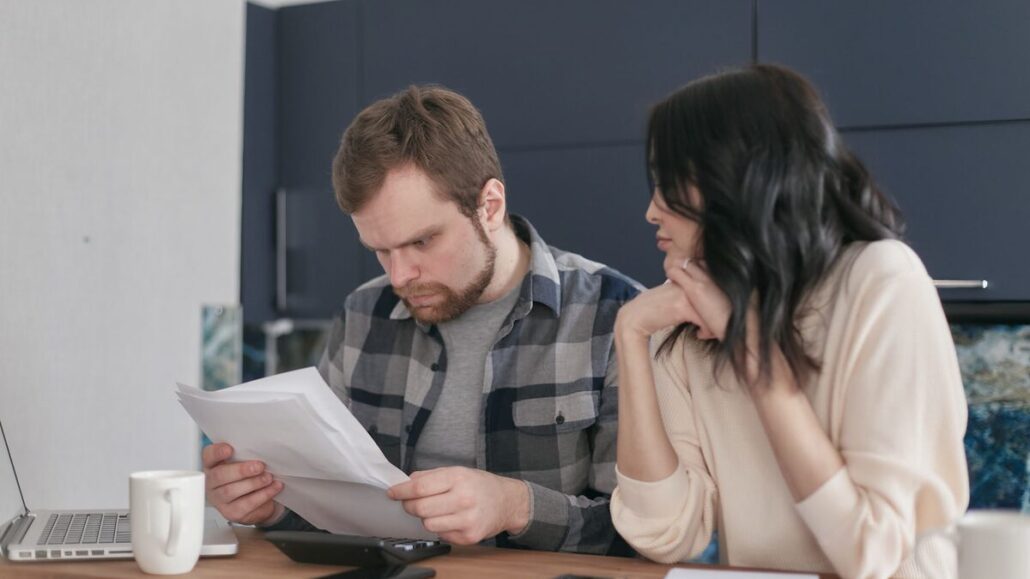 A couple sitting together at a wooden table reading through printed documents, with a laptop and mugs nearby