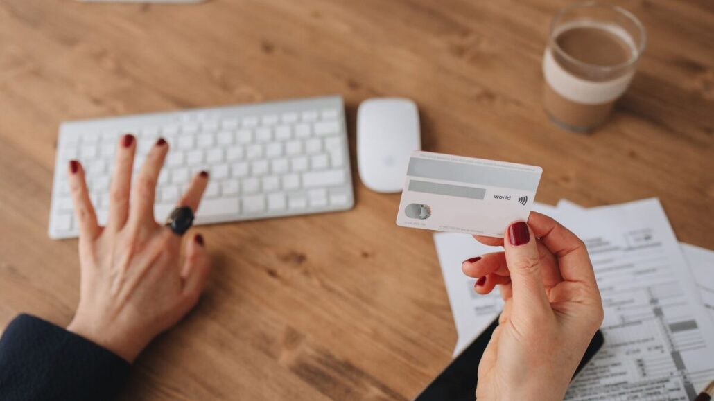 A close-up of hands at a wooden desk, one holding a bank card next to a keyboard, receipts and a glass of coffee