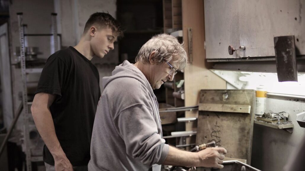 An older craftsman in a grey hoodie showing a younger man how to work a piece of metal in an industrial workshop