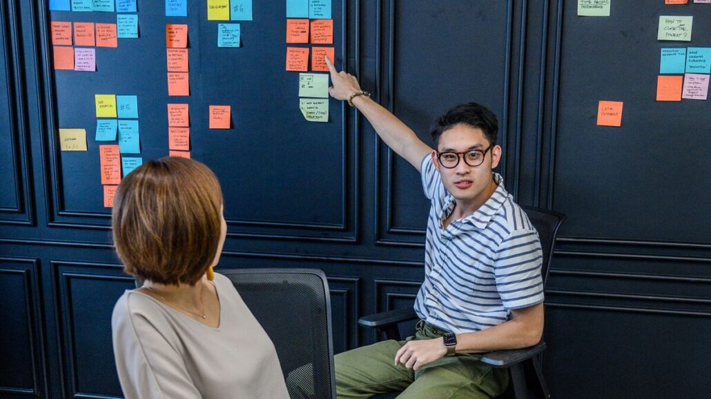 A man in a striped shirt pointing at coloured sticky notes on a dark wall while a colleague listens during a team meeting