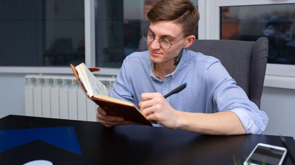 A young man in glasses and a blue shirt writing in a notebook at a dark desk with a phone beside him