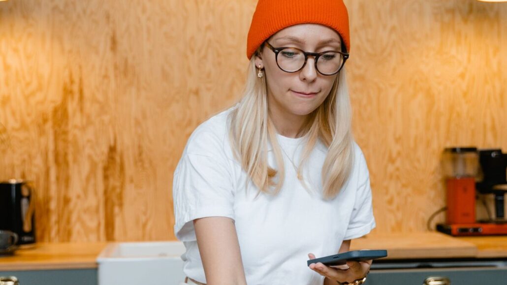 A woman in a beanie and glasses checking her phone at a wooden worktop while packing handmade ceramic dishes