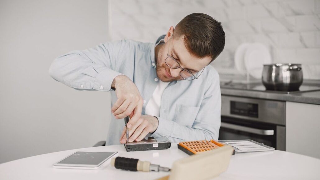 A man in glasses using a screwdriver to repair a small device at a white kitchen table
