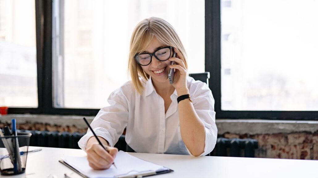 A smiling woman in glasses on a phone call, writing notes on a clipboard at a desk by a large window