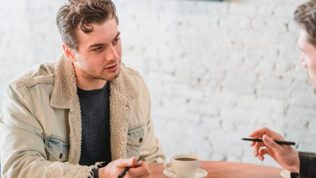 A young man in a sherpa-lined jacket talking with a colleague over coffee and notes at a cafe table against a white brick wall