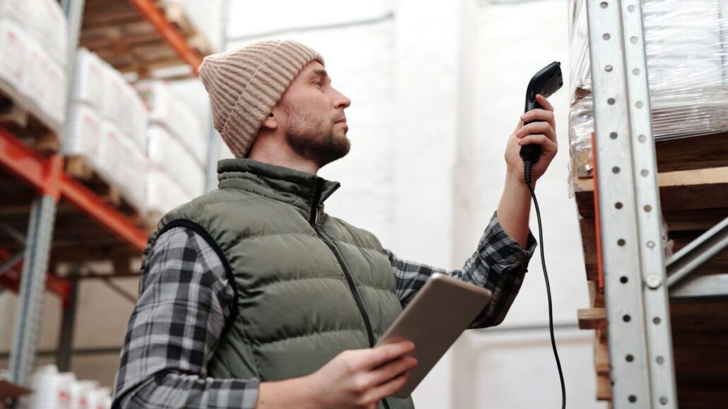 A warehouse worker in a beanie and gilet holding a tablet and a handheld barcode scanner beside tall shelves of stock