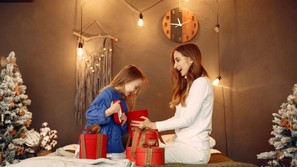 A mother and young daughter opening red gift boxes together in a warmly lit room decorated for Christmas
