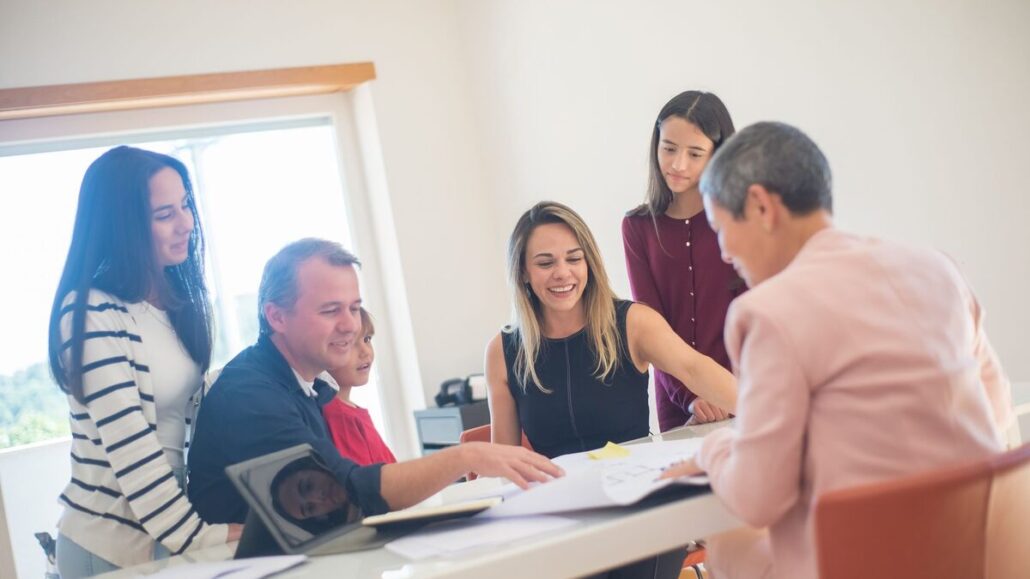 A family of four leaning over a table of papers with an older adviser in a pink jacket in a bright room