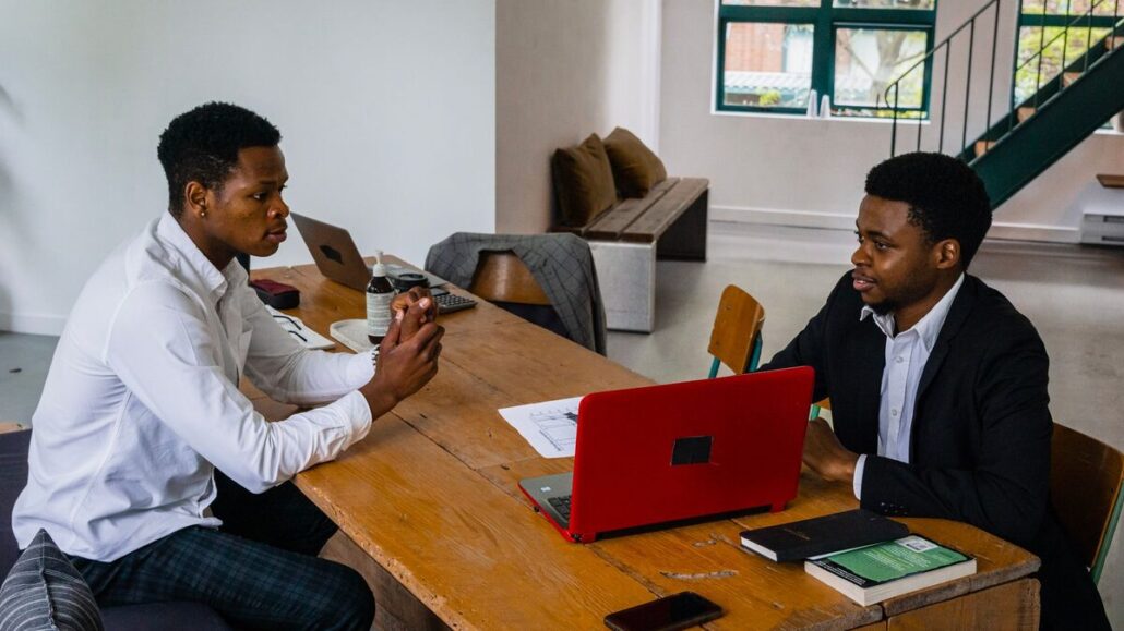 Two men talking across a wooden meeting table with a red laptop, notebook and papers in a bright office